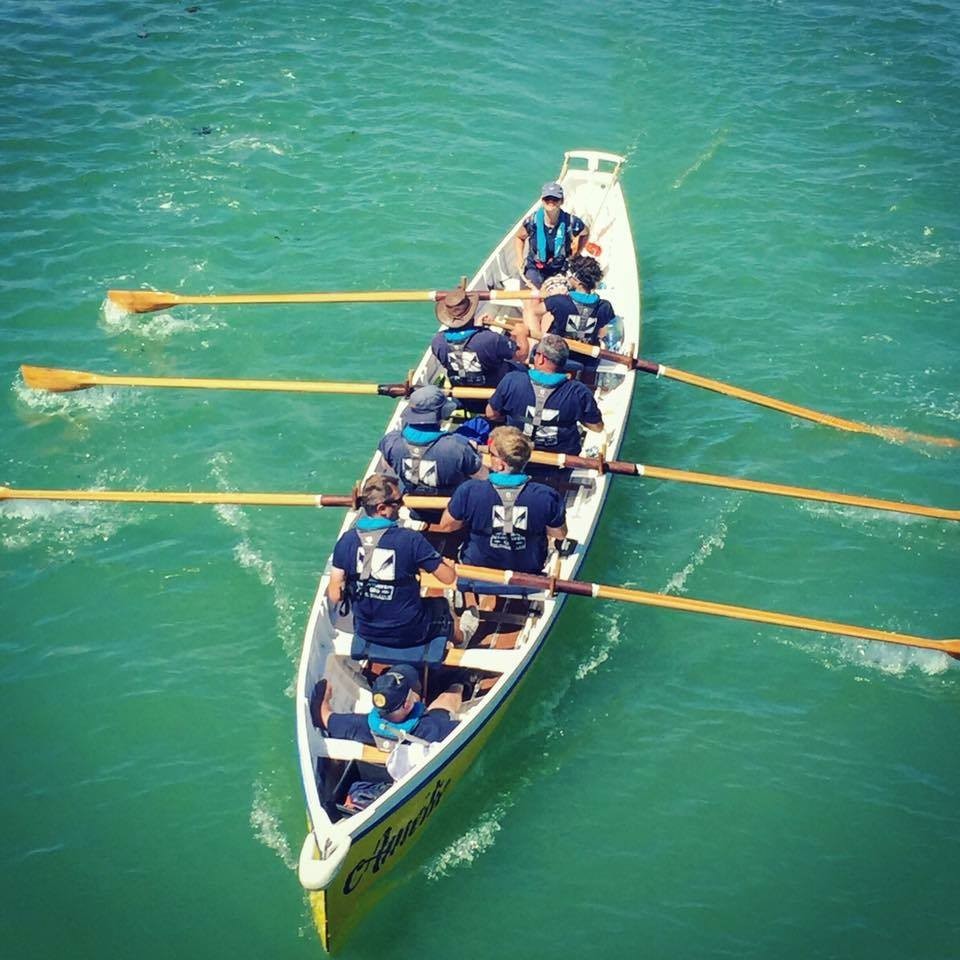 Photo shows people rowing in a gig rowing boat on a bright blue river