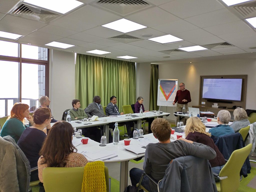 Photo shows a group of people sat around desks listening to a business trainer at the front of the room with a presentation on a screen