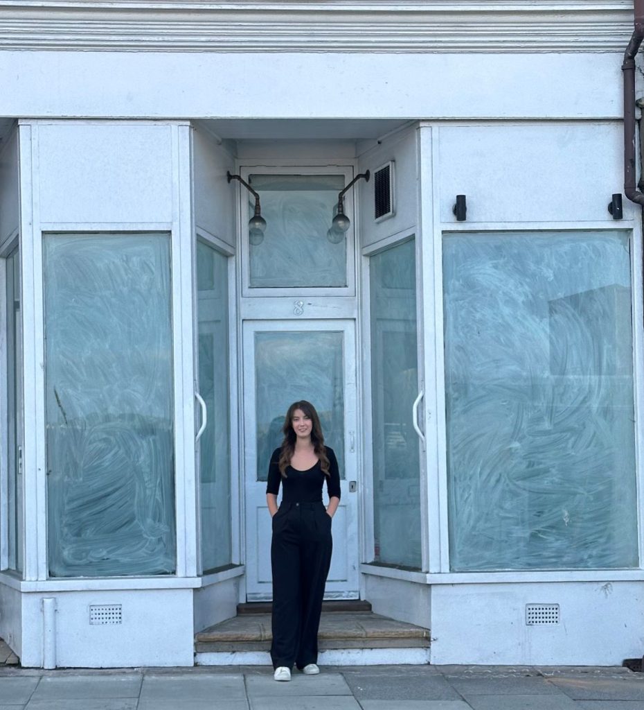 Person in black clothing and white shoes standing on a step in front of a closed storefront with swirled white window coverings, two overhead light fixtures, and the number '8' above the door