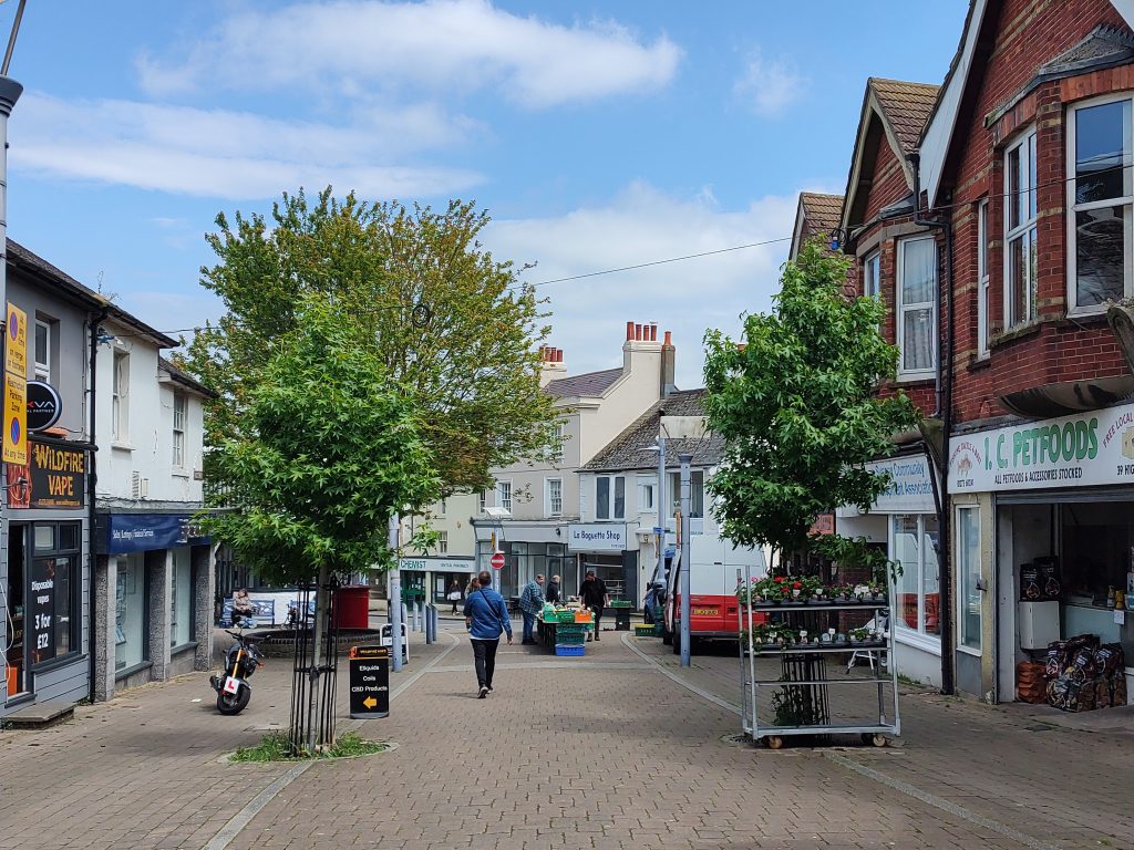 Pedestrian street lined with shops including 'Wildfire Vape' and 'I.C. Petfoods'; two trees in the center, one with a sign for e-liquids and CBD products; a person walking toward another arranging items on display tables under a partly cloudy sky.