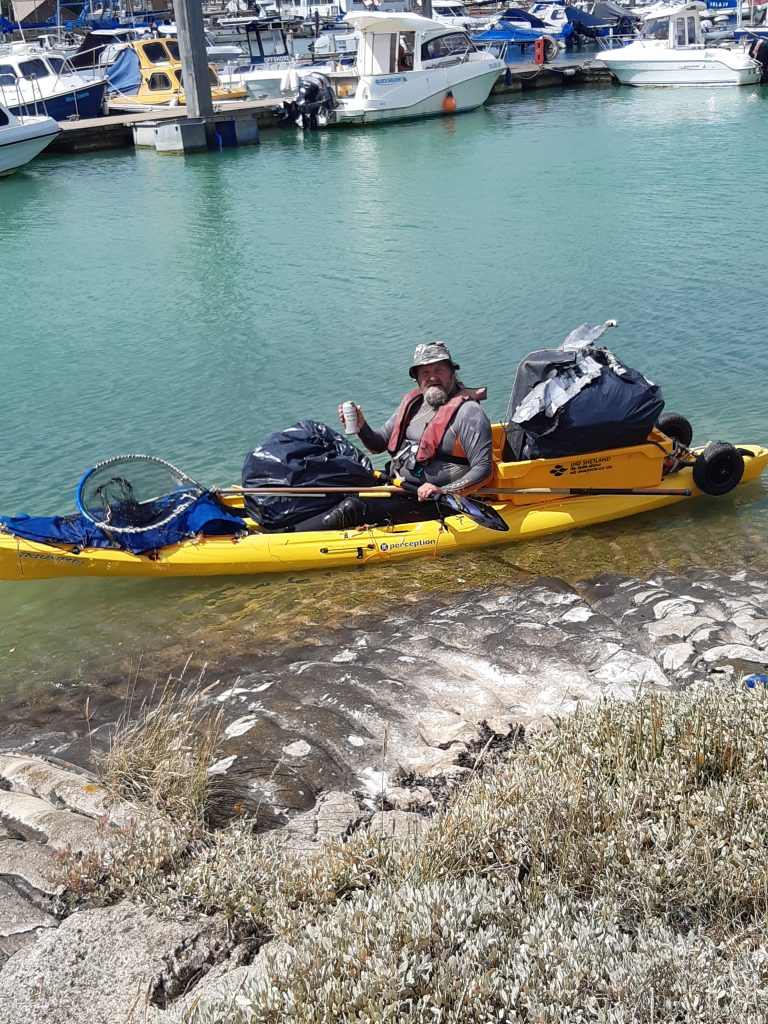 Person in a yellow kayak loaded with gear, docked near a rocky shore in calm greenish-blue water, with other boats visible at a nearby marina