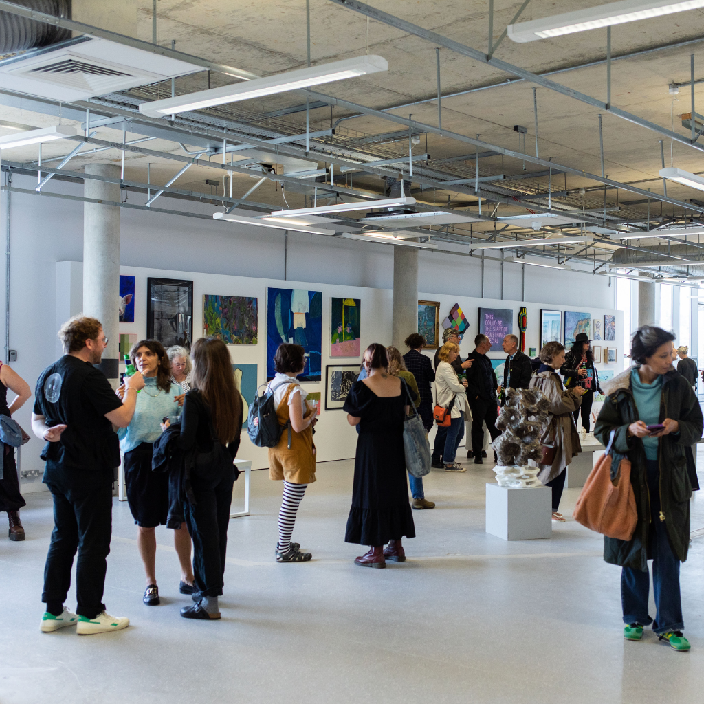 A photo of a large white gallery space hung with a variety of artworks. Lots of people are shown visiting the Newhaven Open Call Exhibition at the BN9 Studio in Newhaven's Marine Workshops.
