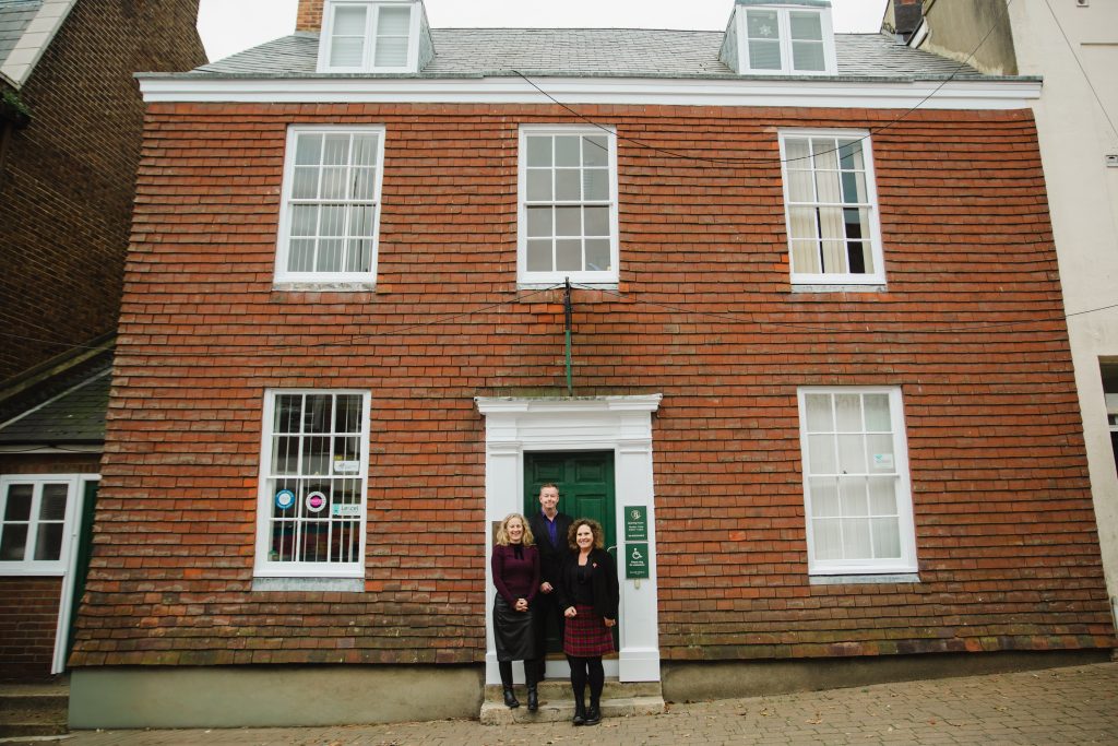Three people standing in front of a two-story brick building with white-framed windows and a green door. The building has a symmetrical design with six large windows and two dormer windows on the roof. There are two green signs mounted next to the entrance