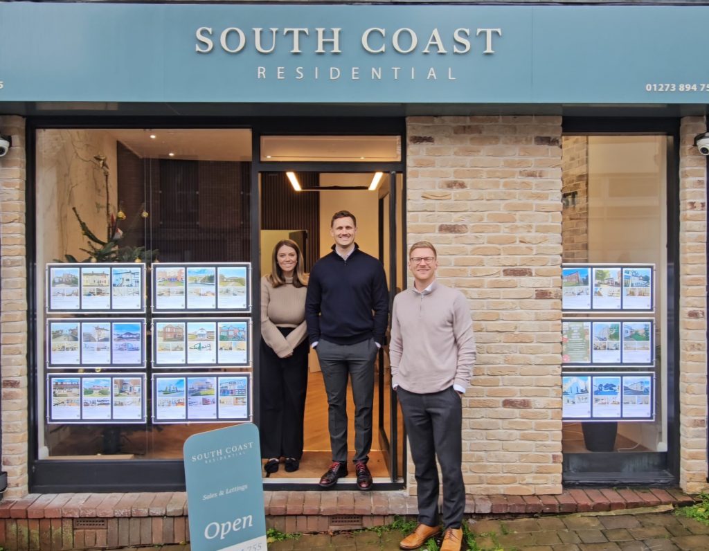 Exterior of South Coast Residential office with a teal sign above the entrance, large windows displaying property listings, and a freestanding sign that reads ‘Open.’ Three people are standing in the doorway.