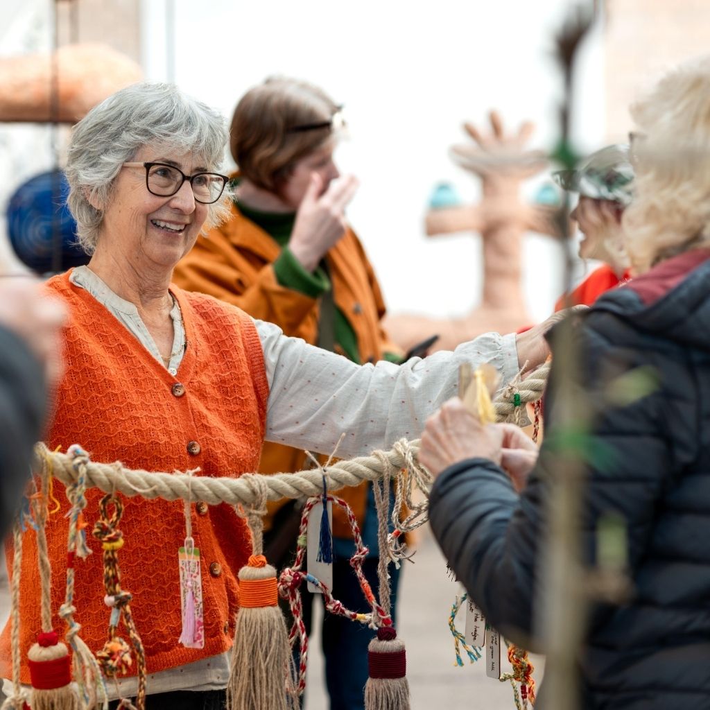 A photograph of a woman with short grey hair and wearing glasses, a long-sleeved top and an orange knitted waitcoast. She holds out a heavy hemp rope strung with brightly coloured tassles to a person standing in front of her.